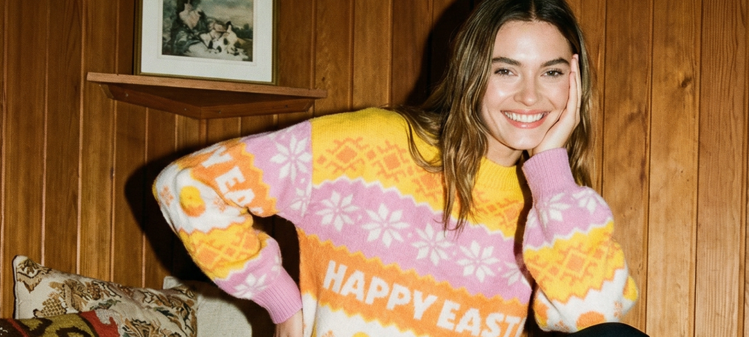 Lächelnde Frau in rosa-gelbem Norwegerpullover mit Happy-Easter-Schriftzug vor Holzwand