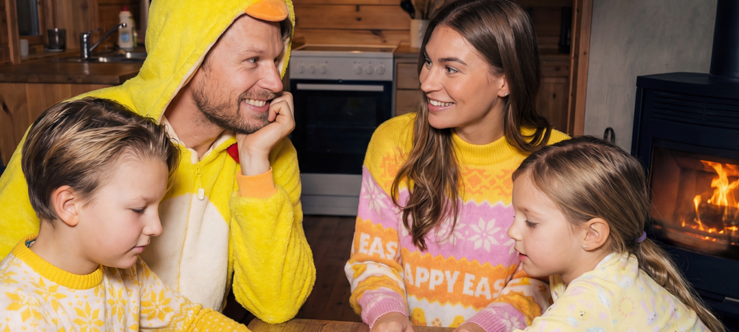 Familie in Osterpullovern und Kostüm sitzt am Tisch vor einem Kamin in gemütlicher Holzküche