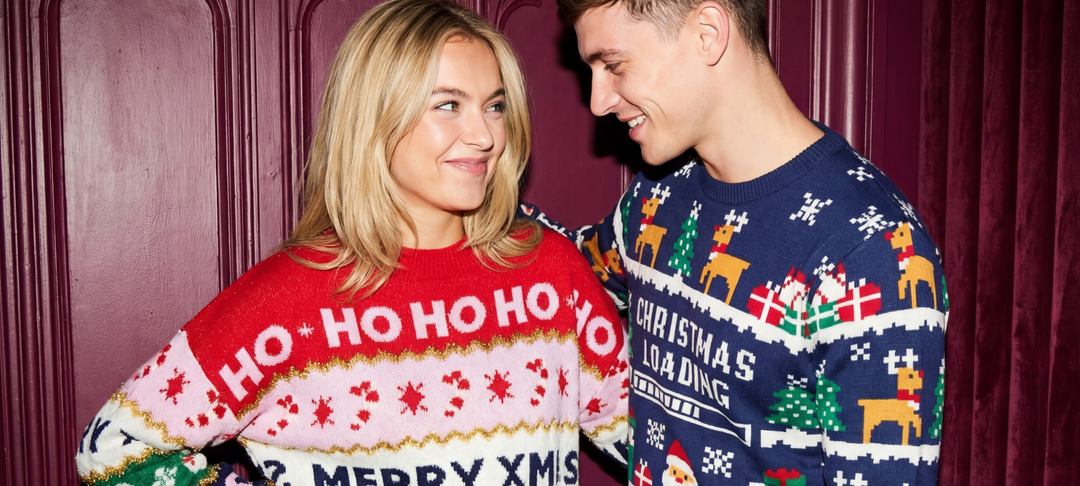 Lächelndes Paar in bunten Weihnachtspullovern mit Ho-Ho-Ho- und Rentier-Motiven vor dunkelrotem Hintergrund