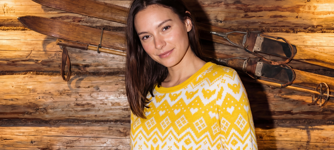 Lächelnde Frau im gelb-weißen Norwegerpullover vor Holzwand mit alten Skiern