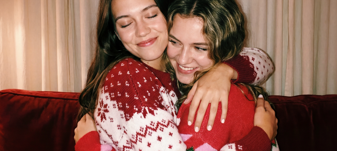 Zwei lächelnde Frauen in roten Weihnachtspullovern umarmen sich auf einem Sofa